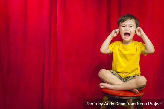 A young boy sitting on a stool with his fingers in his ears in front of a red curtain